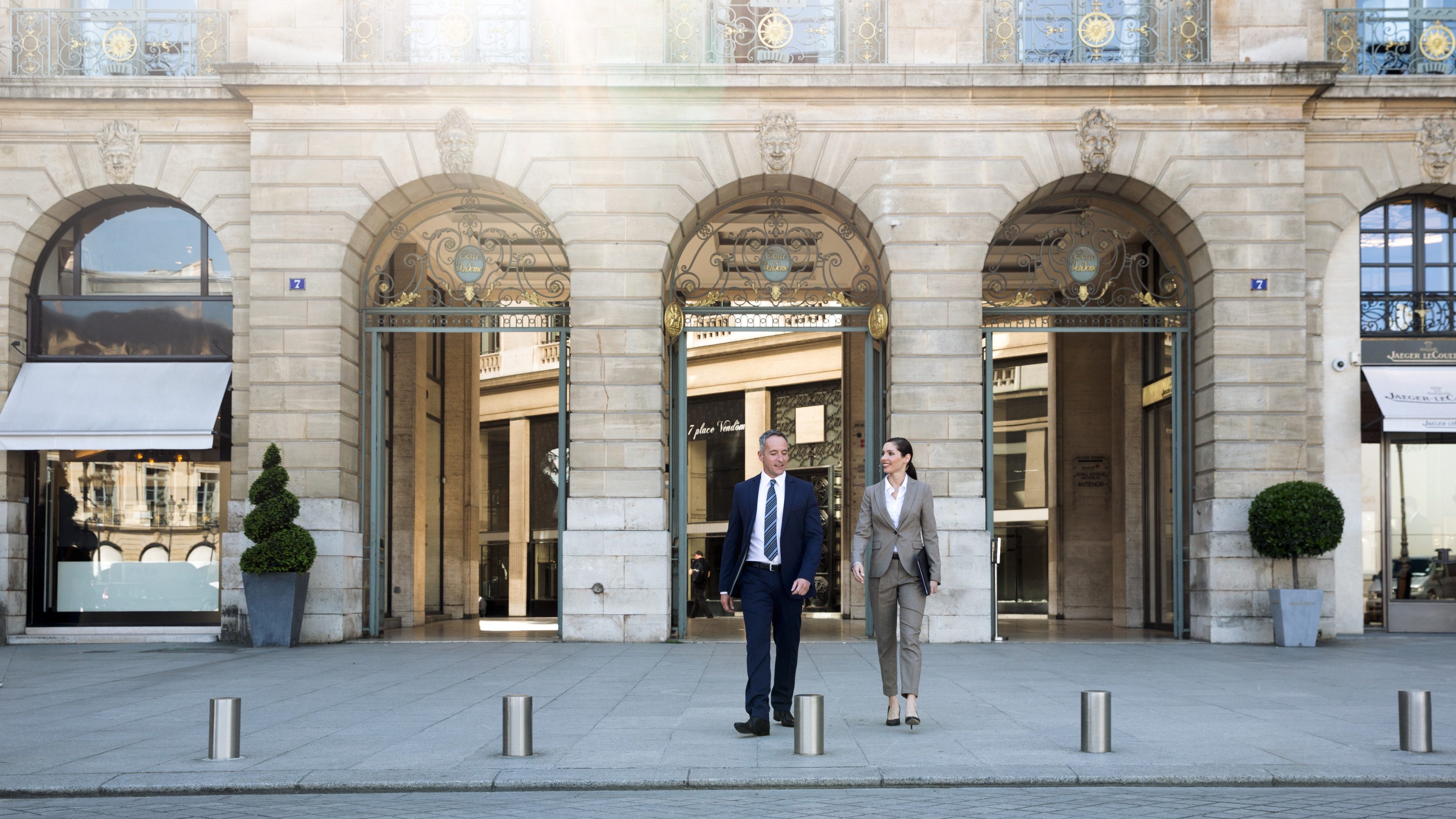 Group of four consisting of two women and two men in business attire on a square in front of a prestigious building façade. The sun’s rays are reflected in one of the windows in the background. Conversation between two men and a woman.