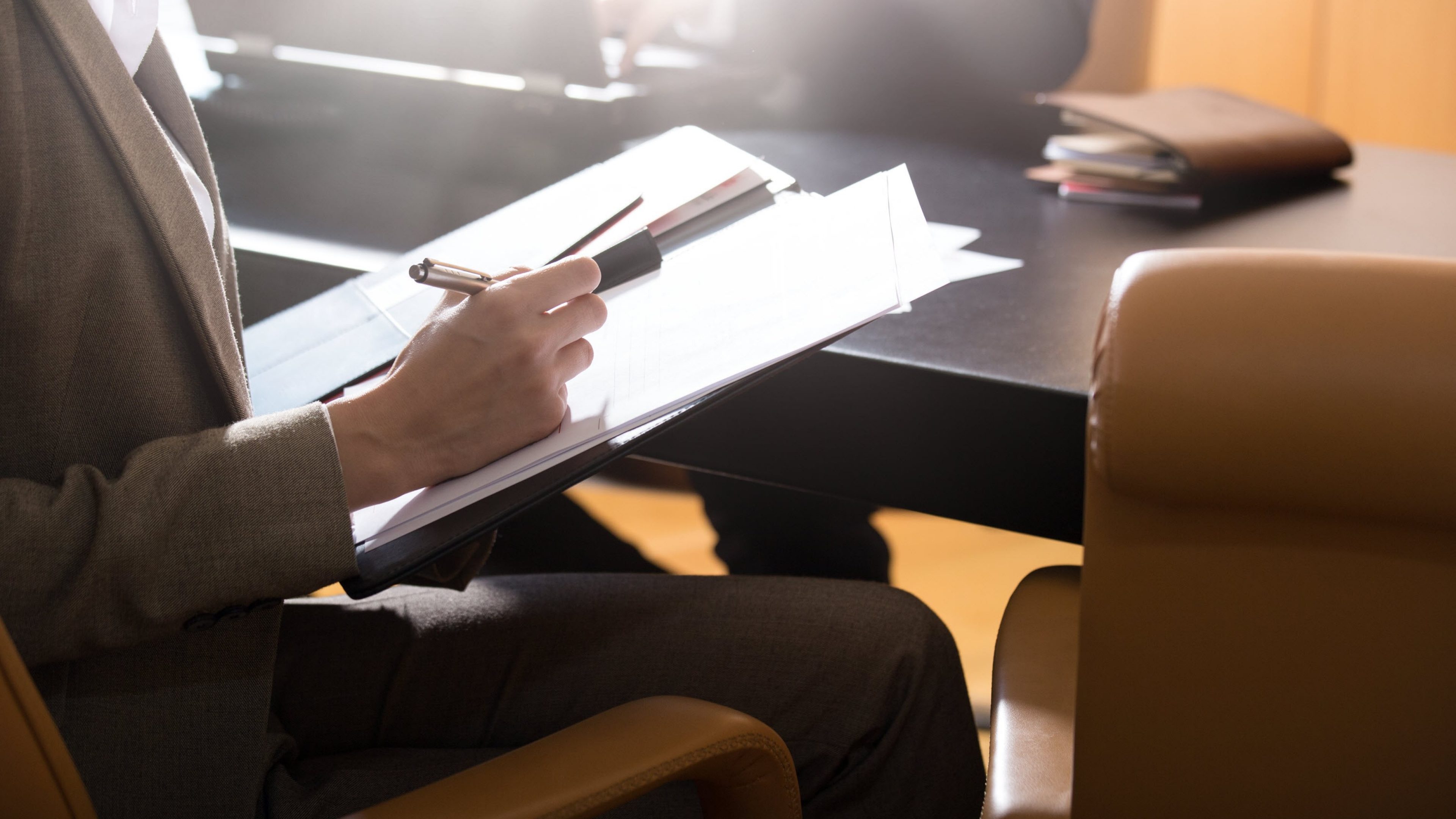 Person sitting at a table with leather armchairs, holding documents and a pen in their hand. In the background is a half-open diary and an open laptop that another person is working on. Sun shines into the picture from behind.