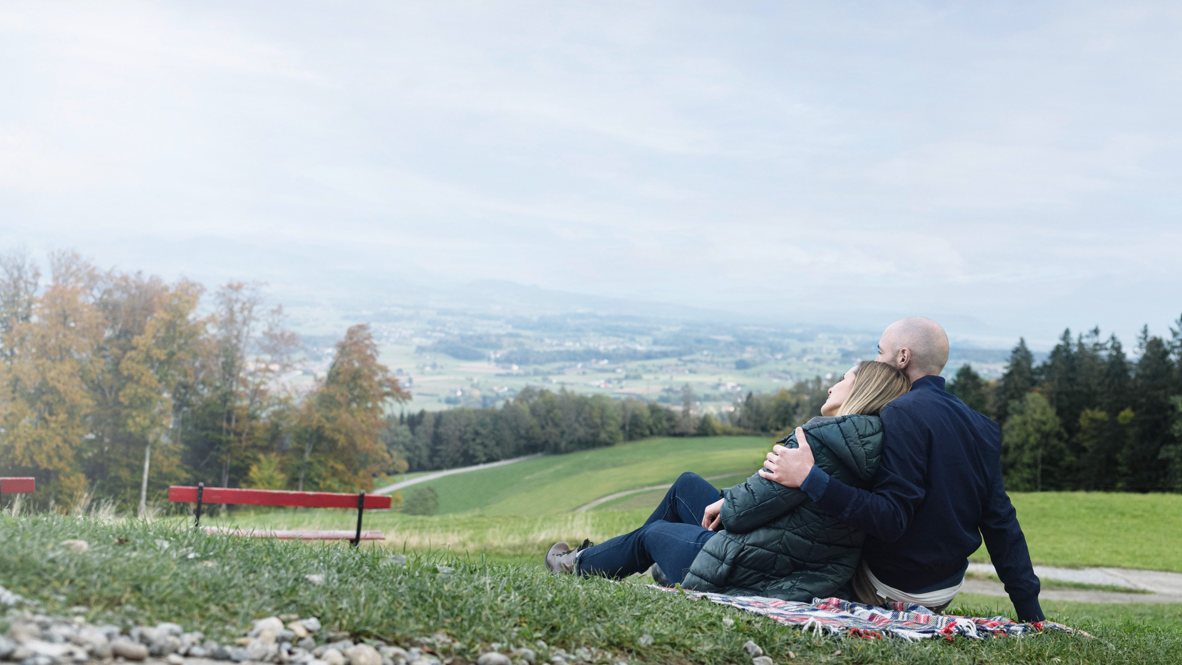 A man and a woman sitting on a blanket by the side of the road. The man has his arm around the woman. They both look at a wooded valley.