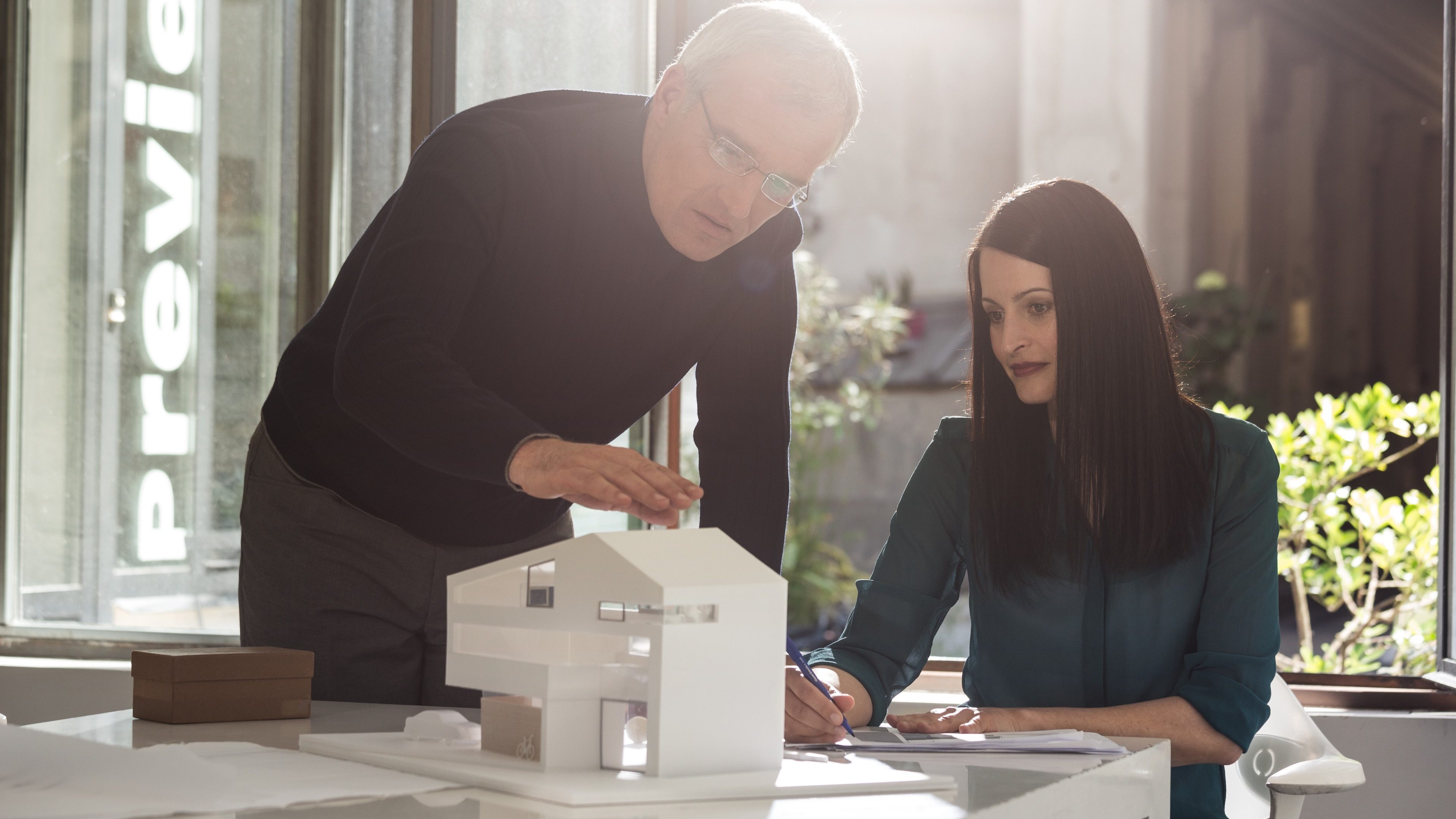 A seated woman and a man standing up and leaning on a desk both looking at a model of a house; sun rays in the background