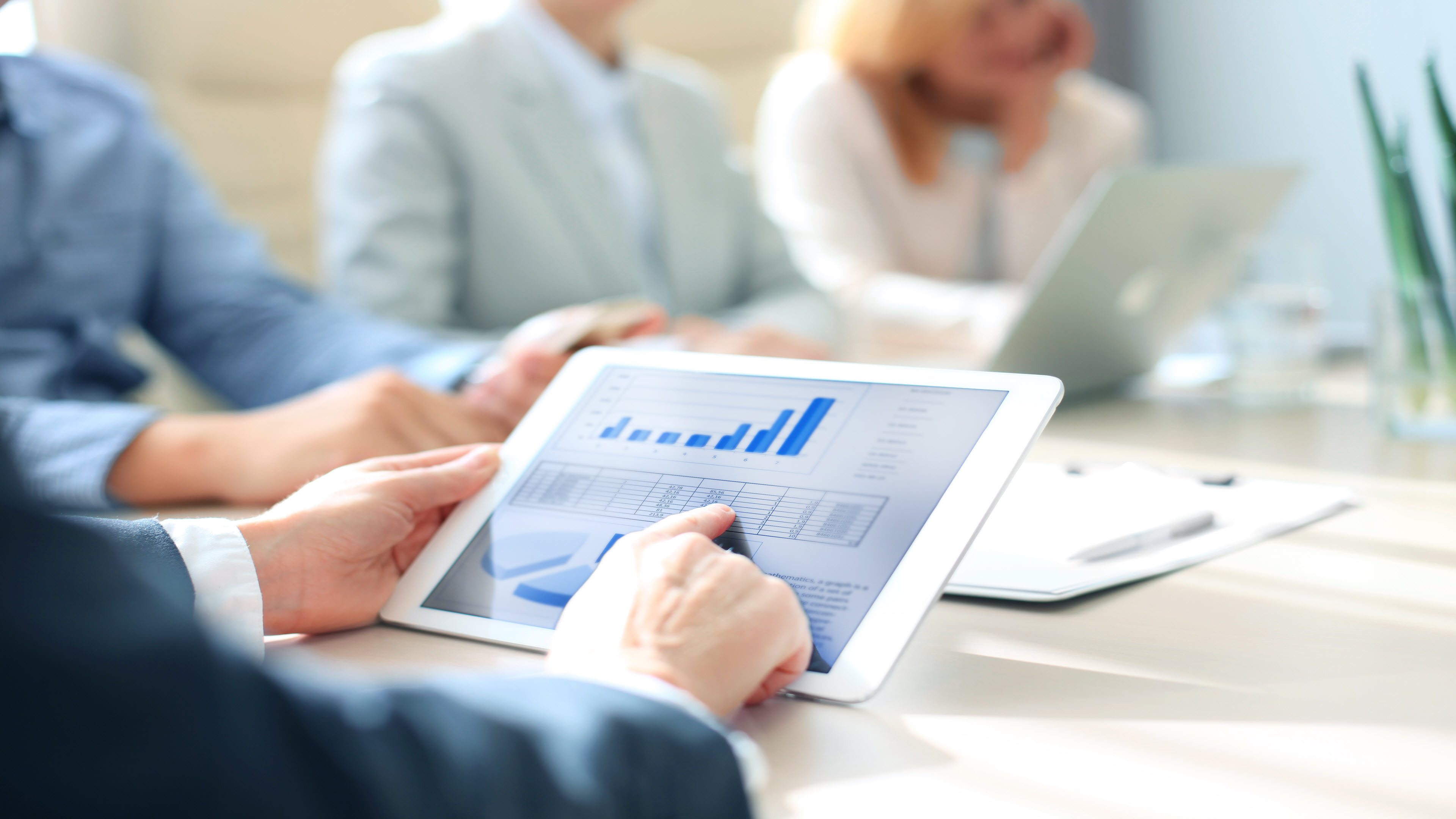 A hand from a man pointing to a tablet on which a graphic about an economy can be seen. Other people are sitting in the background.