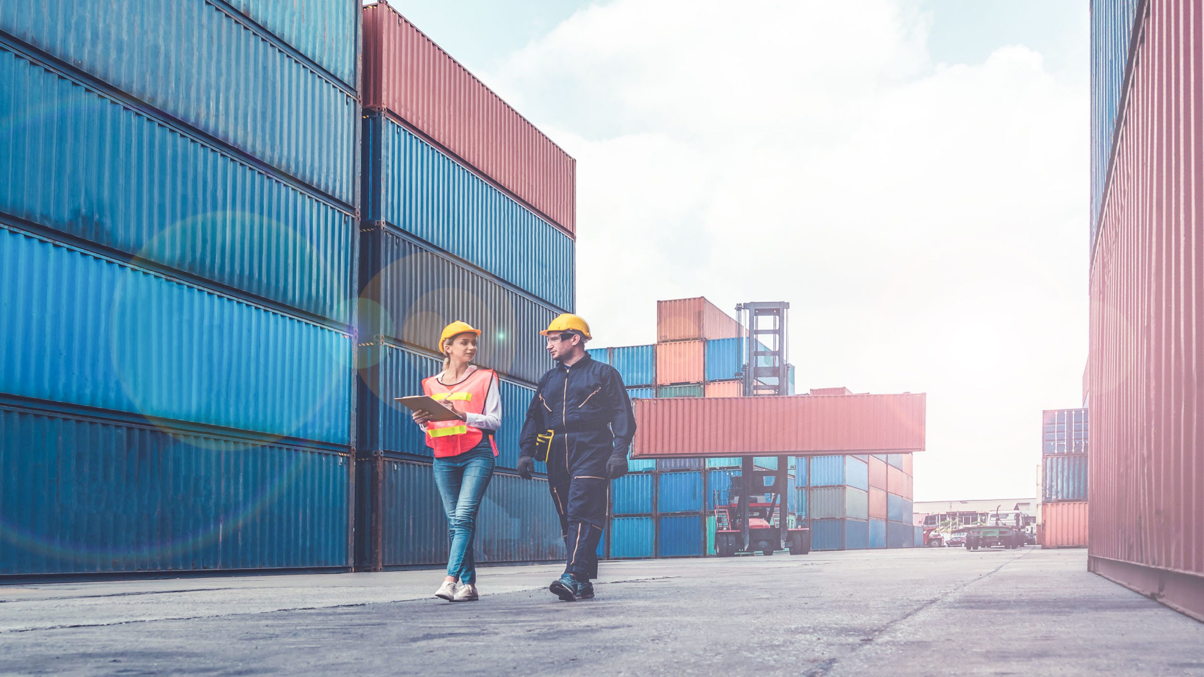 A woman and a man walking through a container harbour. In the background you can see a forklift loading the containers.