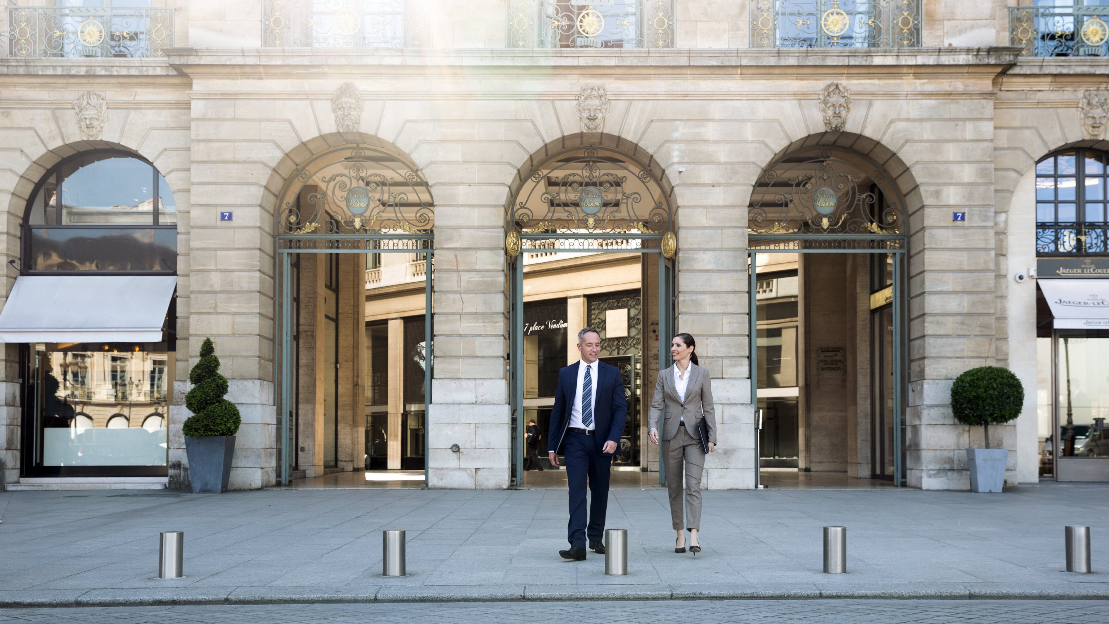 Vierergruppe aus zwei Frauen und zwei Männern in Businesskleidung auf einem Platz vor einer repräsentativen Hausfassade. Sonnenstrahlen reflektieren in einem der Fenster im Hintergrund. Unterhaltung zweier Männer mit einer Frau.