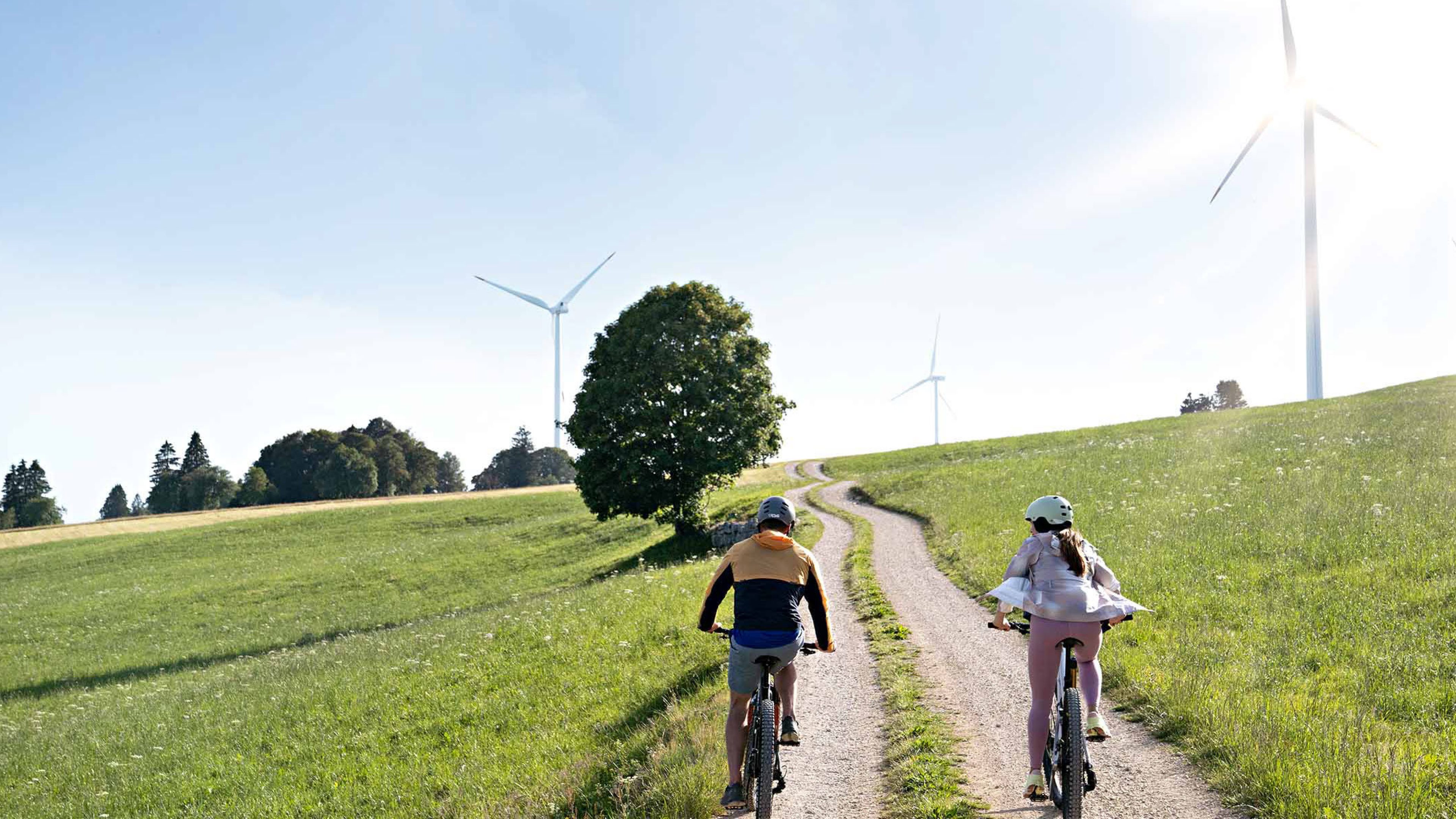 Due persone vanno in bicicletta su una strada sterrata, sullo sfondo si vedono delle pale eoliche.