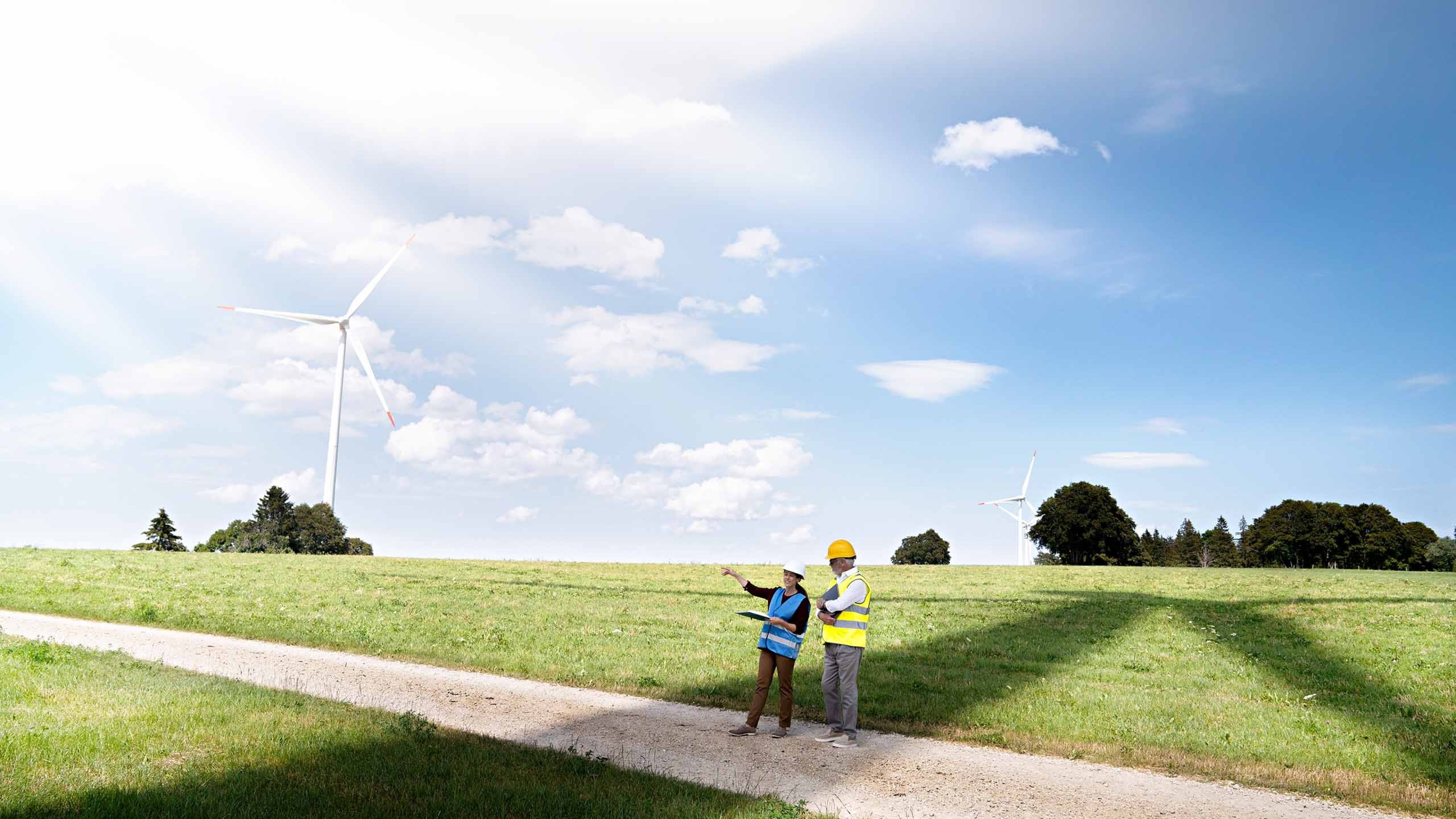 Un campo in cui due persone, che indossano giubbotti catarifrangenti e caschi, progettano le nuove turbine eoliche.